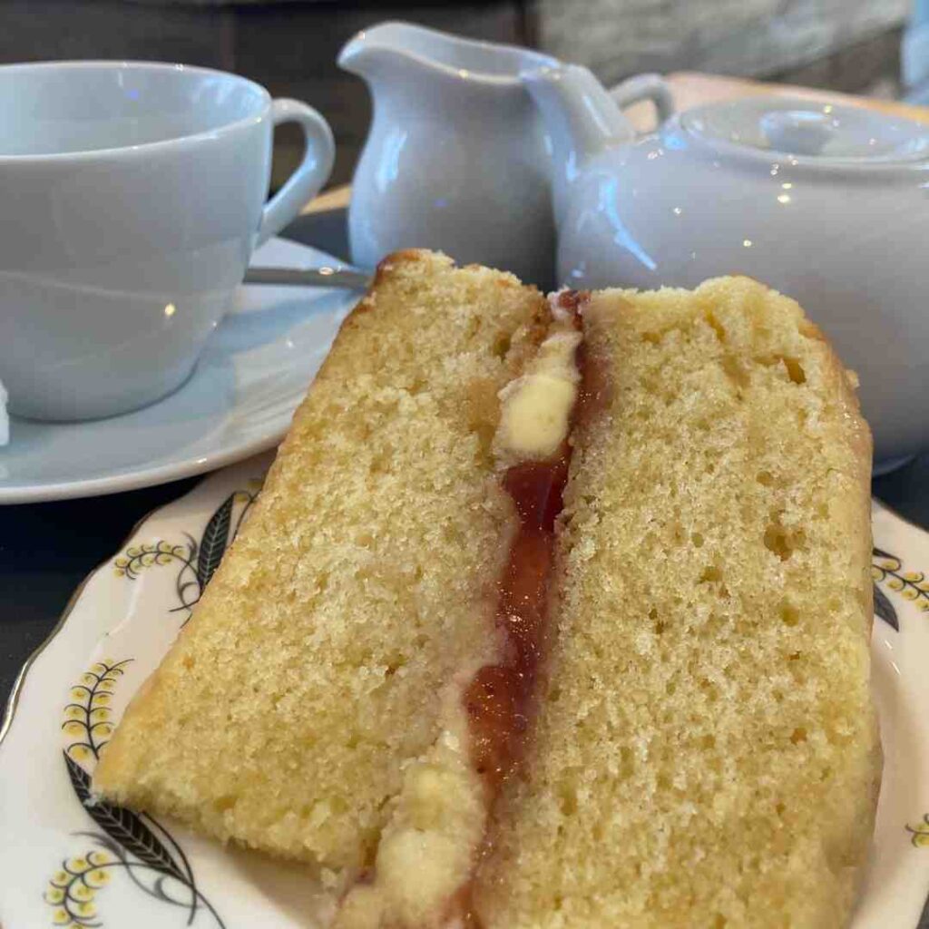 A slice of Victoria sponge cake next to a tea pot, a tea cup and a milk jug
